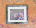 A framed abstract art painting featuring a colorful, textured depiction of a female torso in shades of pink, purple, and blue on a gray background, set in a rustic gray wood frame with a white mat, displayed on a textured orange wall with paint splatters.