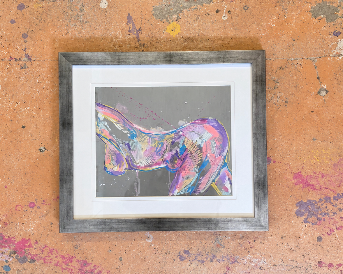 A framed abstract art painting featuring a colorful, textured depiction of a female torso in shades of pink, purple, and blue on a gray background, set in a rustic gray wood frame with a white mat, displayed on a textured orange wall with paint splatters.
